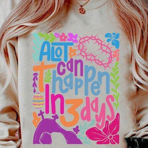 May include: A light beige sweatshirt with a colorful graphic design that says "A lot can happen in 3 days". The design includes flowers, a cross, and a crown of thorns.