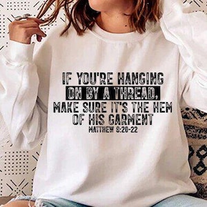 May include: White sweatshirt with the text "IF YOU'RE HANGING ON BY A THREAD, MAKE SURE IT'S THE HEM OF HIS GARMENT" and "MATTHEW 9:20-22" in black. The sweatshirt has long sleeves and a crew neck. The image is of a person wearing the sweatshirt.
