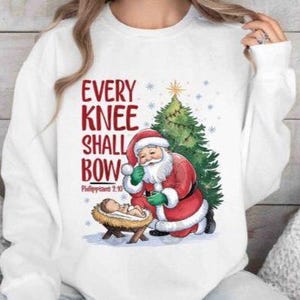 May include: White sweatshirt featuring a Christmas design. The graphic includes Santa Claus, a Christmas tree, and the text "EVERY KNEE SHALL BOW" in red. A baby is in a manger. Snowflakes and a star are also present.