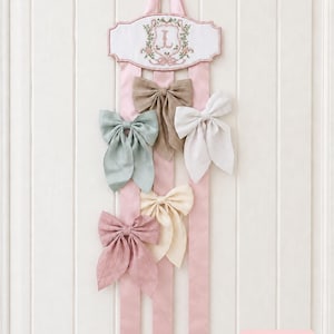 Pode incluir: Um organizador de parede vertical com uma placa de tecido branco apresentando uma letra L rosa bordada rodeada por motivos florais. Três longas fitas de cetim rosa pendem da placa, exibindo cinco laços de tecido em bege, verde sálvia, branco, creme e rosa antigo. Uma pequena etiqueta rosa na parte inferior diz Bows not included.