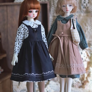 Pode incluir: Duas bonecas vestindo vestidos. A boneca &agrave; esquerda est&aacute; vestindo um vestido preto e branco com uma gola branca e um la&ccedil;o preto. A boneca &agrave; direita est&aacute; vestindo um vestido marrom com uma gola branca e um la&ccedil;o branco. Ambas as bonecas t&ecirc;m cabelo comprido.
