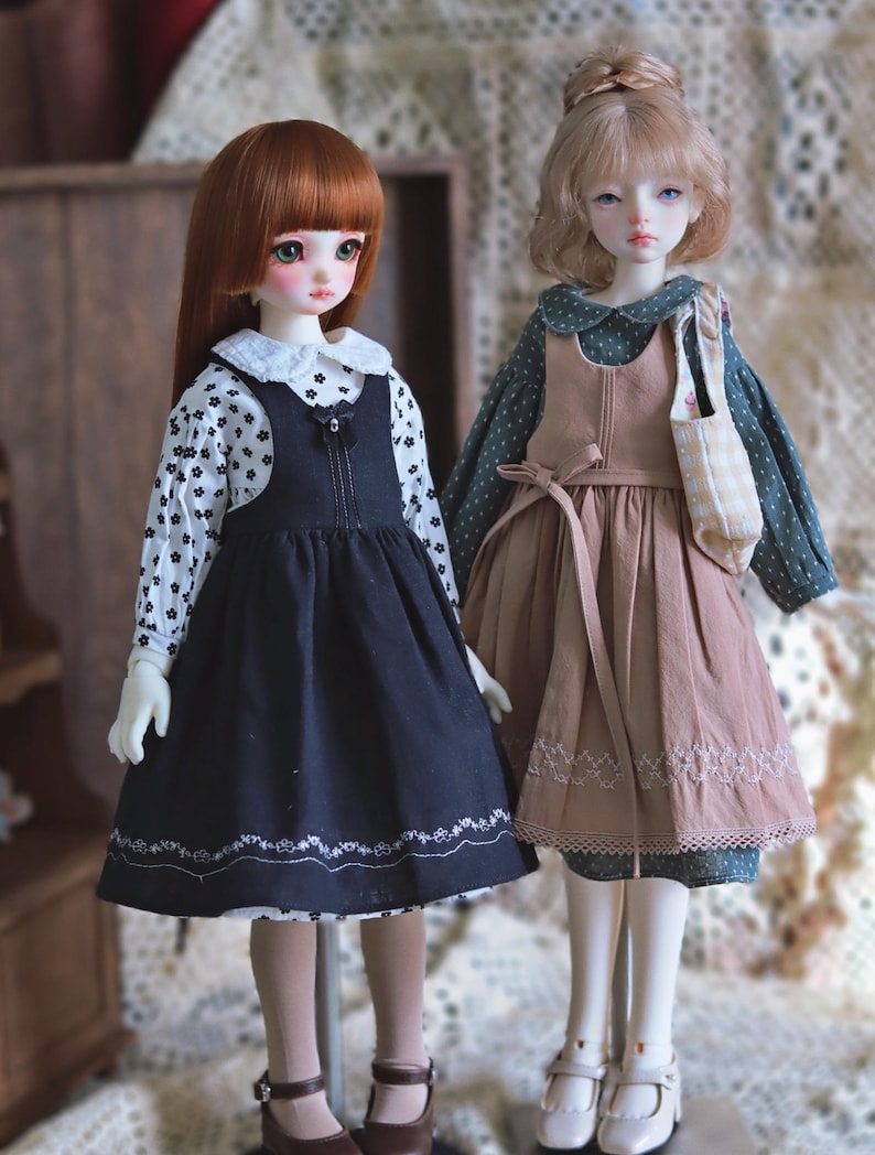 Pode incluir: Duas bonecas vestindo vestidos. A boneca &agrave; esquerda est&aacute; vestindo um vestido preto e branco com uma gola branca e um la&ccedil;o preto. A boneca &agrave; direita est&aacute; vestindo um vestido marrom com uma gola branca e um la&ccedil;o branco. Ambas as bonecas t&ecirc;m cabelo comprido.