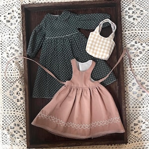 Pode incluir: Dois vestidos de boneca em uma bandeja de madeira. Um vestido &eacute; verde com bolinhas brancas e o outro &eacute; rosa com bordados brancos. Tamb&eacute;m h&aacute; uma pequena bolsa xadrez vichy na bandeja.