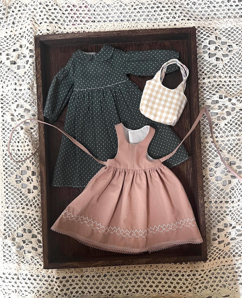 Pode incluir: Dois vestidos de boneca em uma bandeja de madeira. Um vestido &eacute; verde com bolinhas brancas e o outro &eacute; rosa com bordados brancos. Tamb&eacute;m h&aacute; uma pequena bolsa xadrez vichy na bandeja.