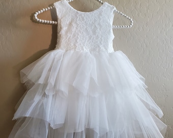 May include: A white sleeveless dress for a young child, hanging on a white beaded hanger. The bodice is made of lace, and the skirt features multiple layers of soft tulle. The dress is designed for special occasions.