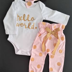 May include: A baby outfit set with a white long-sleeved bodysuit with the text "hello world" in gold glitter, pink pants with gold polka dots, and a pink headband with gold polka dots.