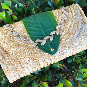 May include: A straw clutch bag with a green leaf design and a rhinestone embellishment. The bag is woven with a light brown and white pattern.