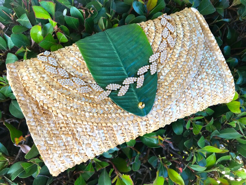 May include: A straw clutch bag with a green leaf design and a rhinestone embellishment. The bag is woven with a light brown and white pattern.