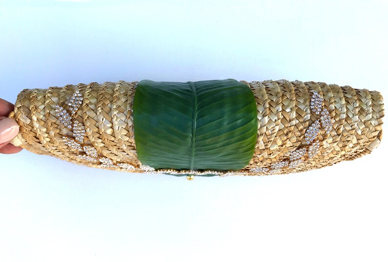 May include: A straw clutch bag with a green leaf design and rhinestones. The bag is woven and has a gold clasp.