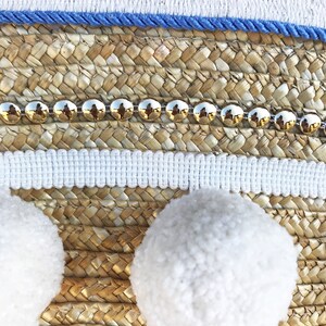 May include: A woven straw tote bag with white pom poms, a blue and white trim, and a row of gold studs.