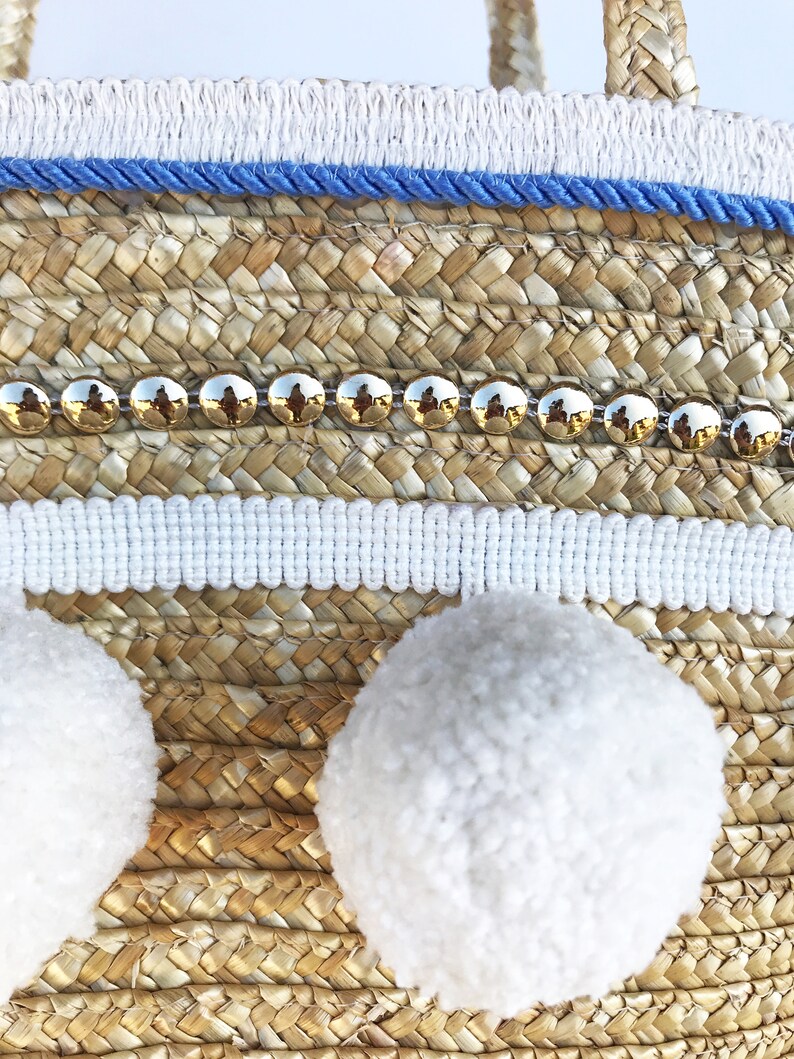 May include: A woven straw tote bag with white pom poms, a blue and white trim, and a row of gold studs.