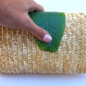 May include: A woven straw clutch bag with a natural beige and white color. The bag is closed and a green leaf is partially covering the top.