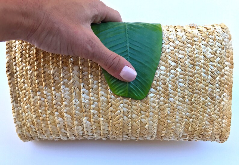 May include: A woven straw clutch bag with a natural beige and white color. The bag is closed and a green leaf is partially covering the top.