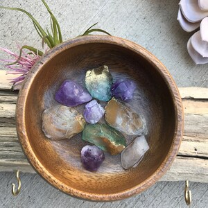 May include: A wooden bowl filled with various crystals, including amethyst, clear quartz, and green aventurine. The bowl is sitting on a wooden surface.