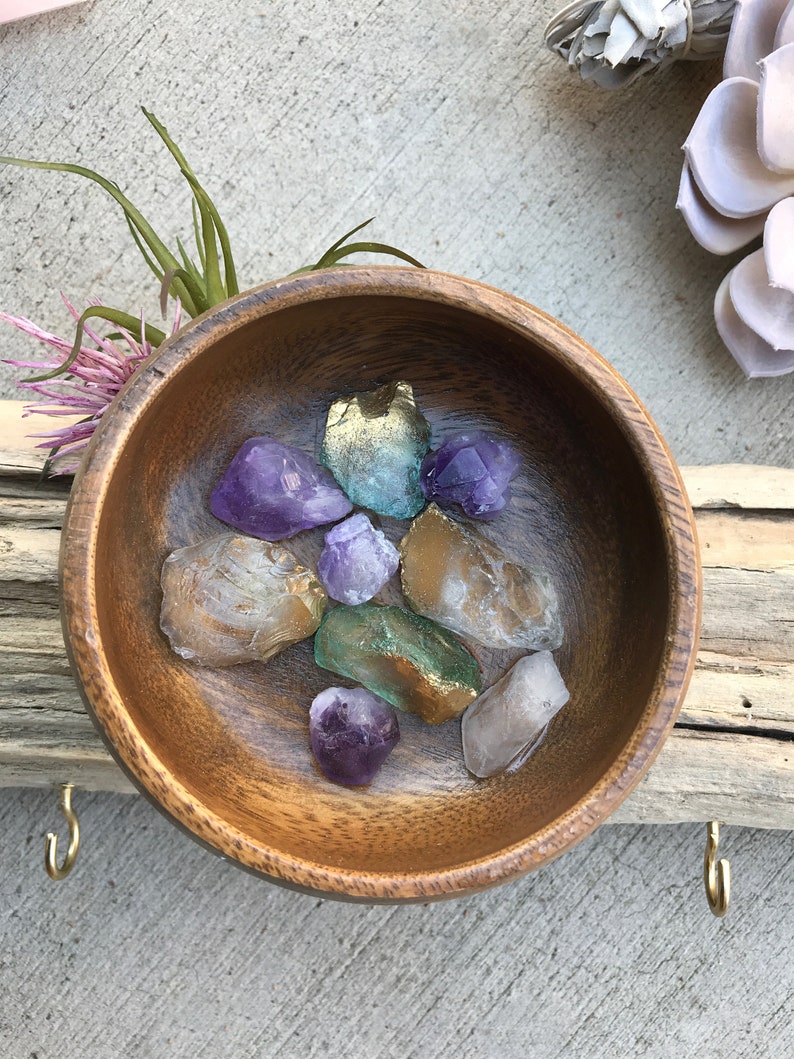 May include: A wooden bowl filled with various crystals, including amethyst, clear quartz, and green aventurine. The bowl is sitting on a wooden surface.