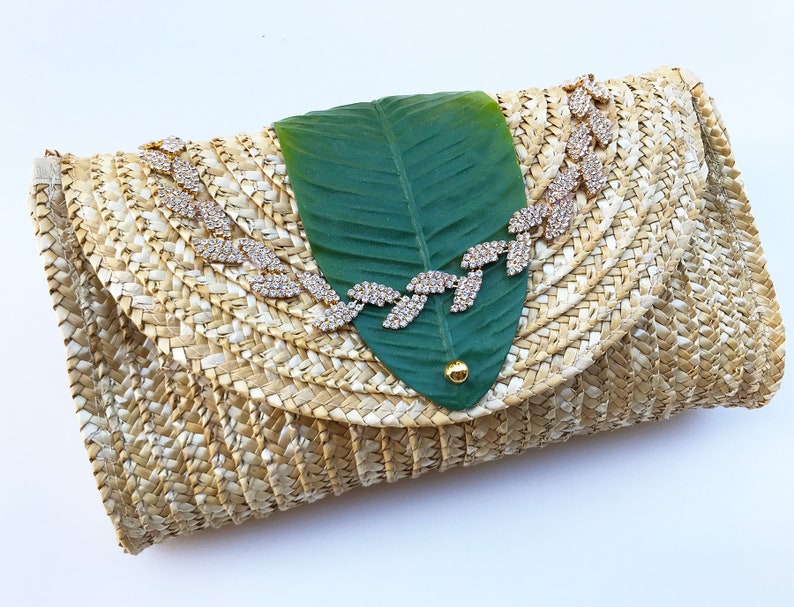 May include: A woven straw clutch bag with a green leaf design and a gold rhinestone embellishment.