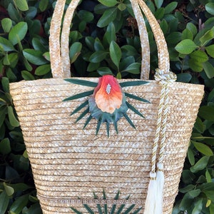 May include: A straw tote bag with a white and gold tassel. The bag is decorated with green leaves and orange flowers.