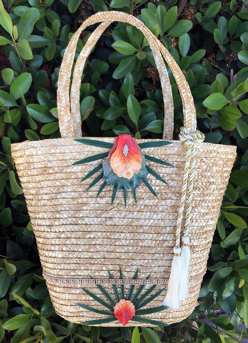 May include: A straw tote bag with a white and gold tassel. The bag is decorated with green leaves and orange flowers.