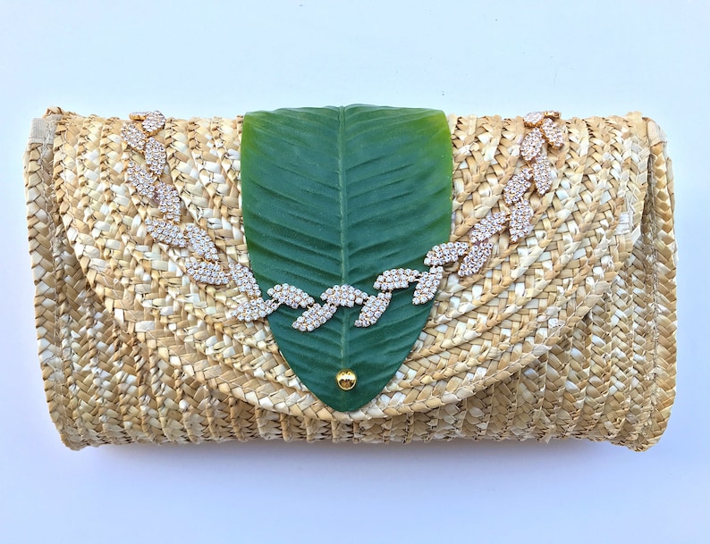 May include: A straw clutch bag with a green leaf design and a rhinestone embellishment.