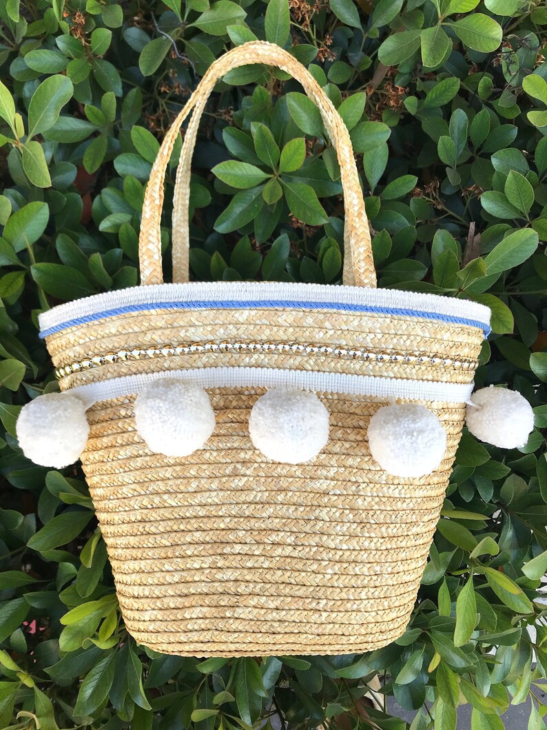 May include: A straw tote bag with white pom poms around the top edge. The bag has a blue and white trim and a gold chain detail.