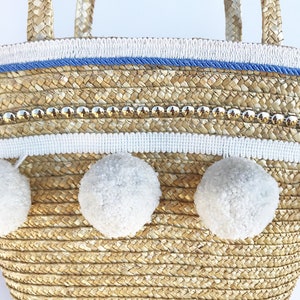 May include: A straw tote bag with white pom poms, a blue and white trim, and gold accents.