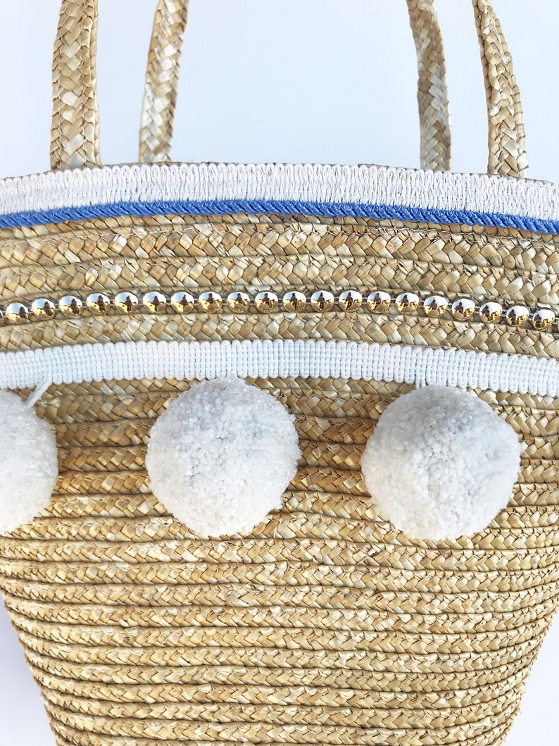 May include: A straw tote bag with white pom poms, a blue and white trim, and gold accents.