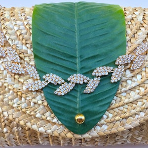 May include: A straw clutch bag with a green leaf design and a rhinestone embellishment. The bag is woven with a natural straw material.