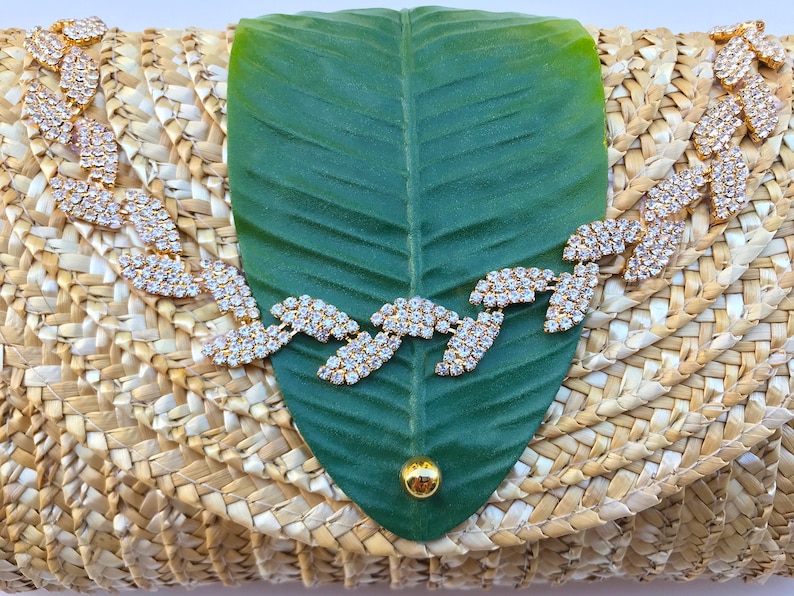 May include: A straw clutch bag with a green leaf design and a rhinestone embellishment. The bag is woven with a natural straw material.