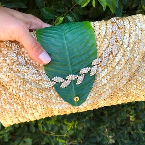 May include: A straw clutch bag with a green leaf design and a rhinestone embellishment. The bag is woven with a light brown straw and has a flap closure.