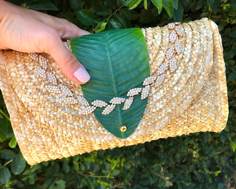May include: A straw clutch bag with a green leaf design and a rhinestone embellishment. The bag is woven with a light brown straw and has a flap closure.