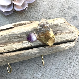 May include: A driftwood key holder with two gold hooks and three crystals. The crystals are a clear quartz, a purple amethyst, and a gold-leafed crystal.