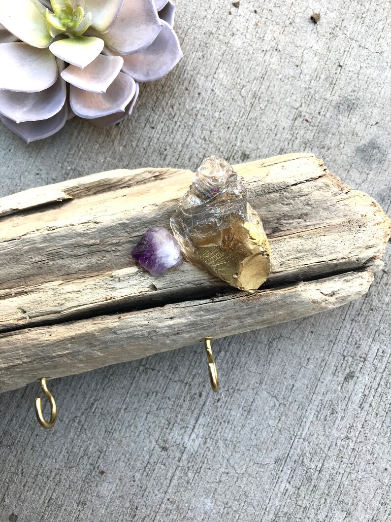 May include: A driftwood key holder with two gold hooks and three crystals. The crystals are a clear quartz, a purple amethyst, and a gold-leafed crystal.