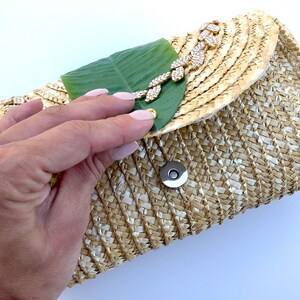 May include: A straw clutch bag with a green leaf and gold rhinestone embellishment. The bag has a woven texture and a magnetic closure.