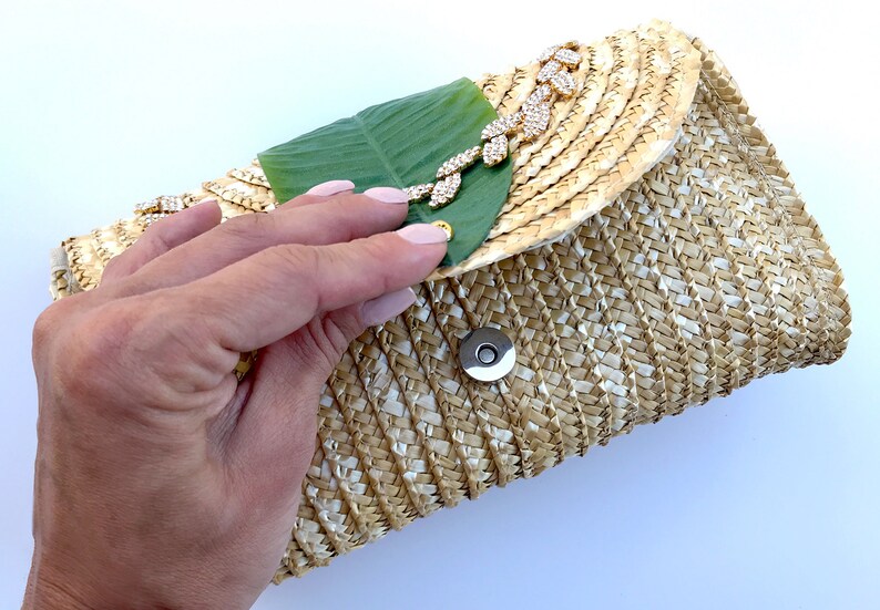 May include: A straw clutch bag with a green leaf and gold rhinestone embellishment. The bag has a woven texture and a magnetic closure.