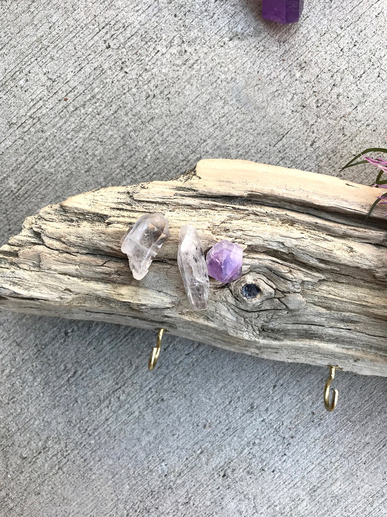 May include: A piece of driftwood with two gold hooks and three crystals: a clear quartz point, a clear quartz cluster, and a purple amethyst.
