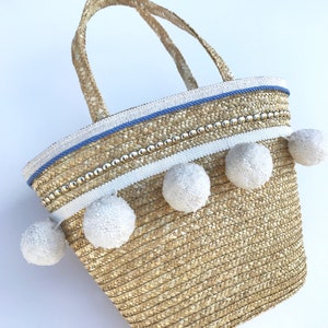May include: A straw tote bag with white pom poms and a blue and white trim. The bag has a beaded detail and two handles.