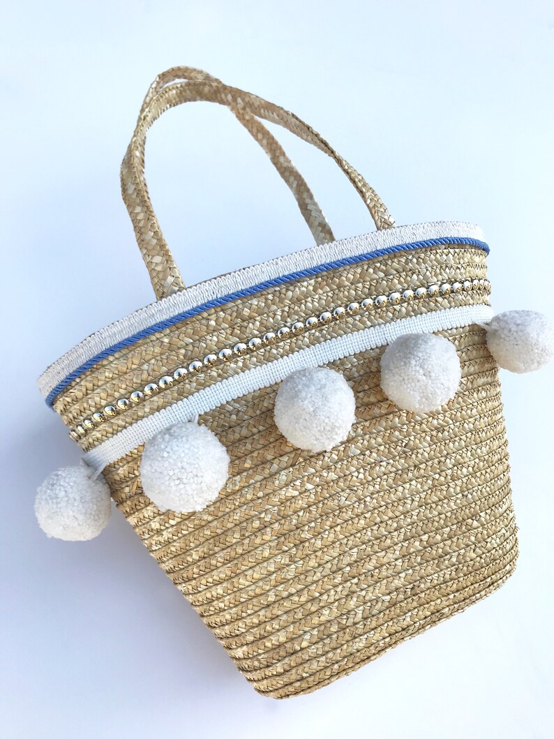 May include: A straw tote bag with white pom poms and a blue and white trim. The bag has a beaded detail and two handles.
