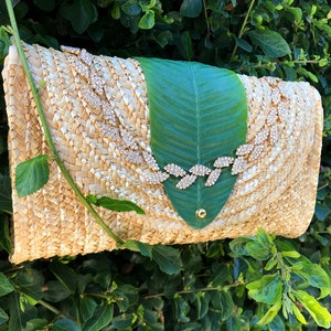 May include: A straw clutch bag with a green leaf design and a gold rhinestone embellishment.