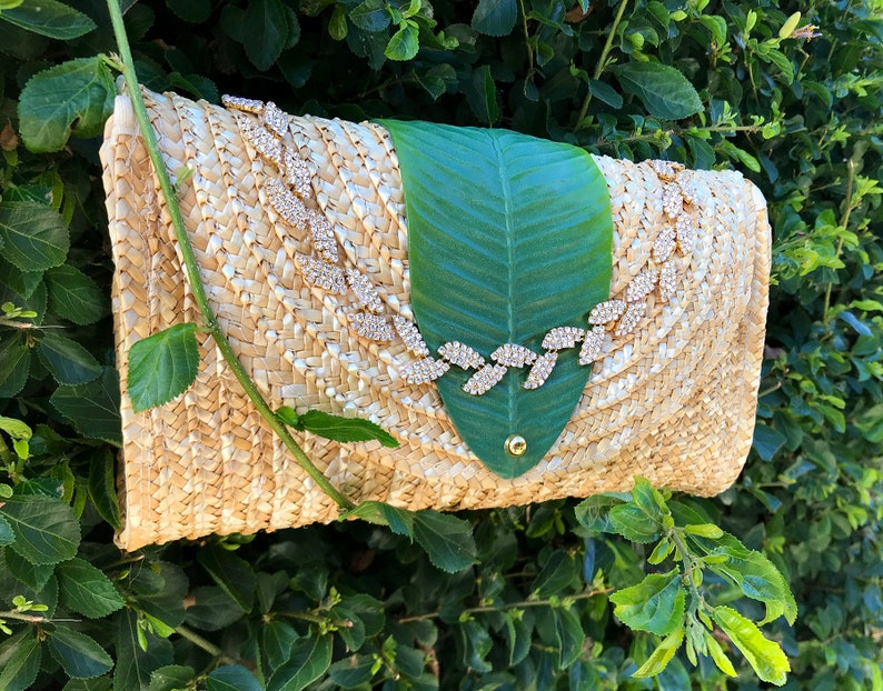 May include: A straw clutch bag with a green leaf design and a gold rhinestone embellishment.