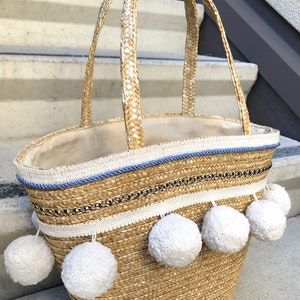 May include: A straw tote bag with white pom poms and blue and white trim. The bag has a woven texture and is decorated with a gold chain.