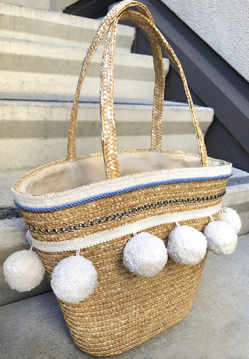 May include: A straw tote bag with white pom poms and blue and white trim. The bag has a woven texture and is decorated with a gold chain.