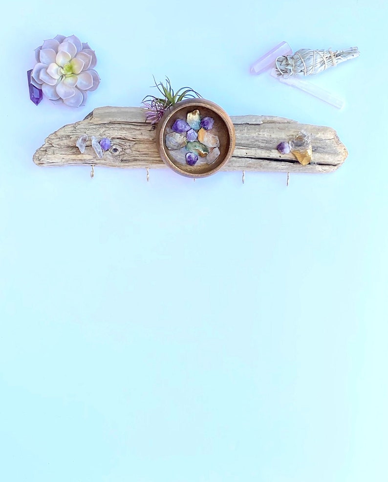 May include: A driftwood key holder with three hooks and a small wooden bowl filled with colourful crystals. A succulent plant is growing out of the bowl. There is a small piece of dried sage and a pink crystal on the white background.