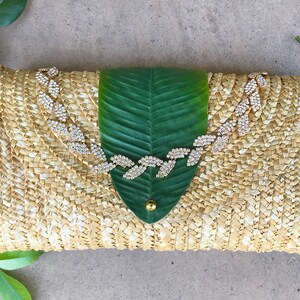 May include: A straw clutch bag with a green leaf design and a rhinestone embellishment. The bag is woven with a light brown and white pattern.