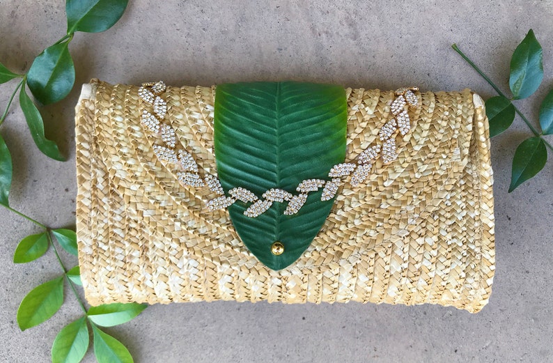 May include: A straw clutch bag with a green leaf design and a rhinestone embellishment. The bag is woven with a light brown and white pattern.