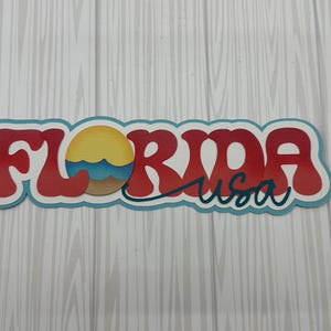 May include: A red and blue decorative sign with the word "FLORIDA" in a retro font. A stylized sun with waves and sand is included in the design. The text "usa" is written in cursive below the word "FLORIDA".