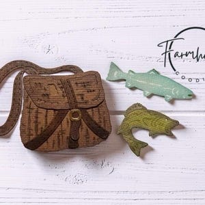 May include: A brown wooden satchel with a strap and buckle detail, accompanied by two wooden fish cutouts, one light blue and one olive green. The text Farmhouse Goodies is printed in black in the upper right corner of the composition.