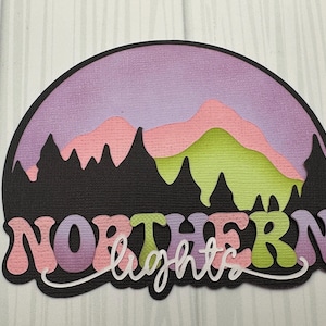 May include: A layered paper cut design featuring a mountain scene under a purple sky. The word "NORTHERN" is in pink, green, and purple, with "lights" in white script below. The design is on a black background.