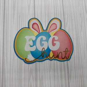 May include: A colorful Easter-themed cutout featuring three decorated eggs in green, blue, and pink. The word "EGG" is in white, and "hunt" is in gold script. Bunny ears are at the top.