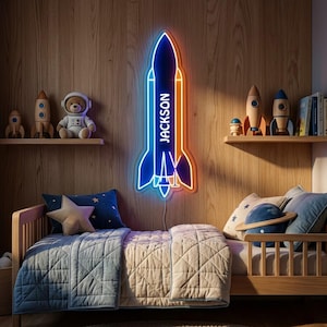 Könnte beinhalten: Ein raketenförmiges Neonschild mit dem Namen JACKSON in weißer Schrift. Das Schild leuchtet blau und orange und ist an einer holzverkleideten Wand über einem Holzbett mit blauer Steppdecke, sternförmigem Kissen und planetenförmigem Kissen montiert.