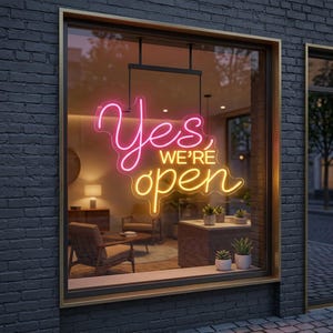 May include: A neon sign mounted in a storefront window displays the text Yes WE'RE open in glowing pink and yellow script lettering. The sign is suspended against a dark gray brick exterior wall, illuminating the interior view of a room.